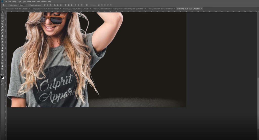 Una donna con lunghi capelli biondi in un'onda sciolta, che indossa occhiali da sole e una maglietta grigia con la scritta Culprit Apparel. Sta sorridendo con un braccio alzato, su uno sfondo nero con un'interfaccia di editing digitale dove i suoi capelli vengono scontornare capelli in Photoshop.
