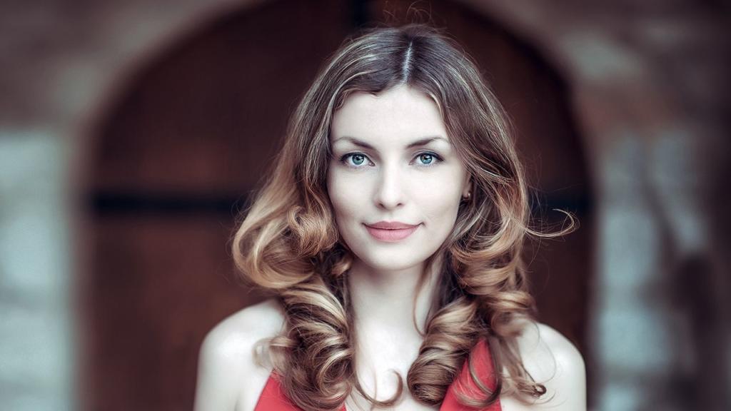 Una persona con lunghi capelli ricci e un top rosso è in piedi all'aperto davanti a una porta ad arco, il suo sorriso gentile catturato in modo impeccabile. È come se scontornare i capelli in Photoshop non fosse necessario per evidenziare la loro bellezza naturale.