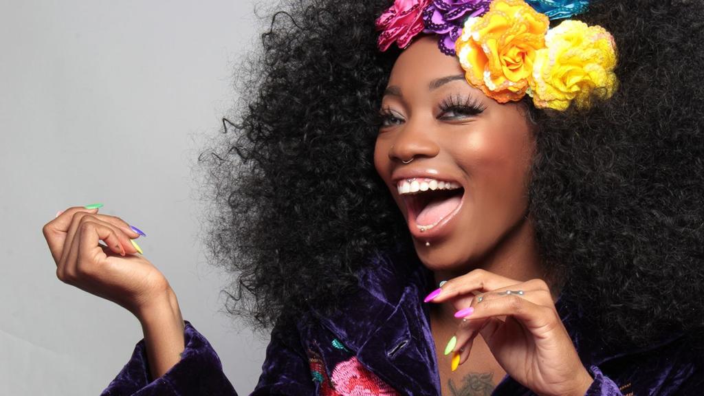 Una donna con i capelli neri ricci e fiori colorati sorride ampiamente. Indossa una giacca viola con motivi floreali e mostra con orgoglio le sue unghie luminose e multicolori. La sua espressione vibrante, sapientemente esaltata come un effetto scontornare capelli Photoshop, trasmette gioia e sicurezza.