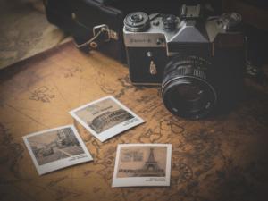 Una macchina fotografica d'epoca è appoggiata su una superficie a tema mappa accanto a tre foto in tonalità seppia, migliorate con una sottile vignettatura in Photoshop. Le immagini presentano punti di riferimento come la Torre Eiffel. La macchina fotografica Zenit-E è appoggiata accanto a una vecchia custodia in pelle, evocando nostalgia.