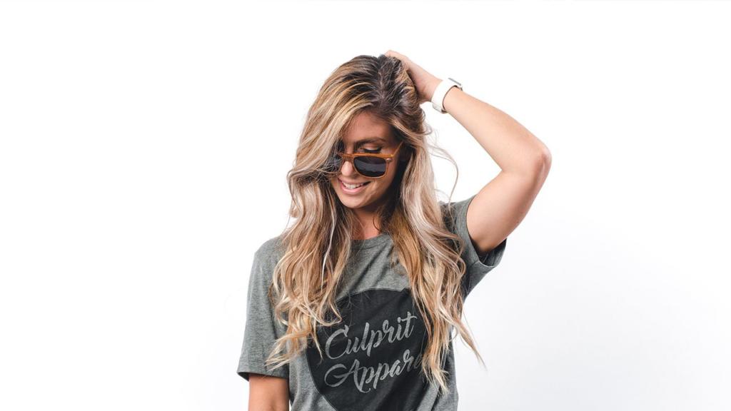 Una donna con lunghi capelli biondi ondulati sorride mentre guarda in basso. Indossa occhiali da sole, una maglietta grigia con una scritta e si tocca giocosamente i capelli su uno sfondo bianco semplice, perfetto per scontornare capelli in Photoshop.