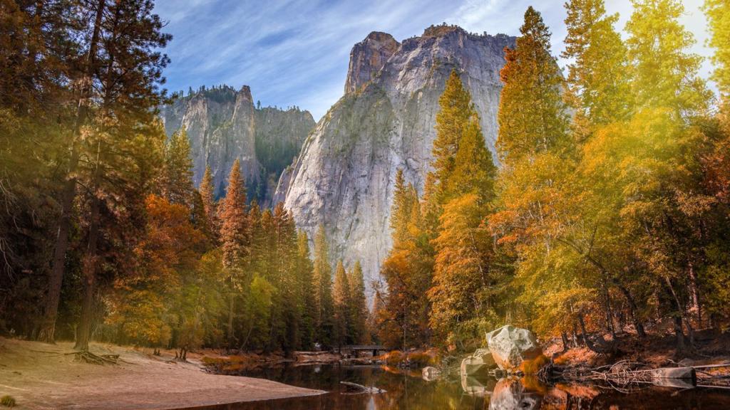 Una vista panoramica di un fiume che riflette gli alti pini circostanti con fogliame autunnale, che ricorda le azioni in Photoshop. Maestose scogliere rocciose si ergono sullo sfondo sotto un cielo azzurro, immerse nella calda luce del sole.