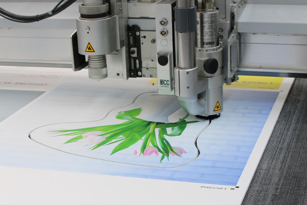 Una macchina da taglio di precisione esegue con perizia il taglio del contorno, tagliando un foglio stampato decorato con motivi a tulipano su uno sfondo blu. Le lame della macchina sono in vista, manovrando senza problemi sulla superficie piana.