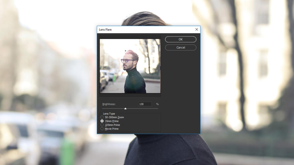 Uno schermo di modifica delle immagini digitali mostra un effetto riflesso lente applicato. Dietro la casella delle impostazioni, un uomo con occhiali da sole e abiti scuri passeggia lungo una strada soleggiata con alberi ed edifici sfocati ad arte sullo sfondo.