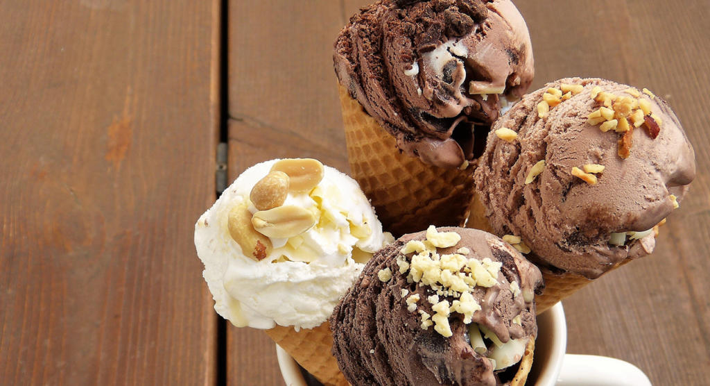 Quattro coni gelato con gusti diversi, tra cui cioccolato e vaniglia, ricoperti di noci, sono sistemati in una coppa. Lo sfondo del tavolo in legno conferisce un'atmosfera rustica, perfetta per chiunque voglia attenuare i bordi con Photoshop per una finitura fotografica impeccabile.