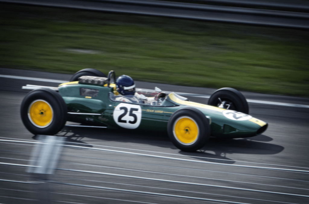 Un'auto da corsa di Formula 1 verde d'epoca con ruote gialle e il numero 25, guidata da un pilota con casco, sfreccia lungo una pista da corsa. Lo sfondo, che ricorda la sfocatura di Photoshop, mostra una vista sfocata della pista e dell'erba verde, catturando l'essenza del movimento.