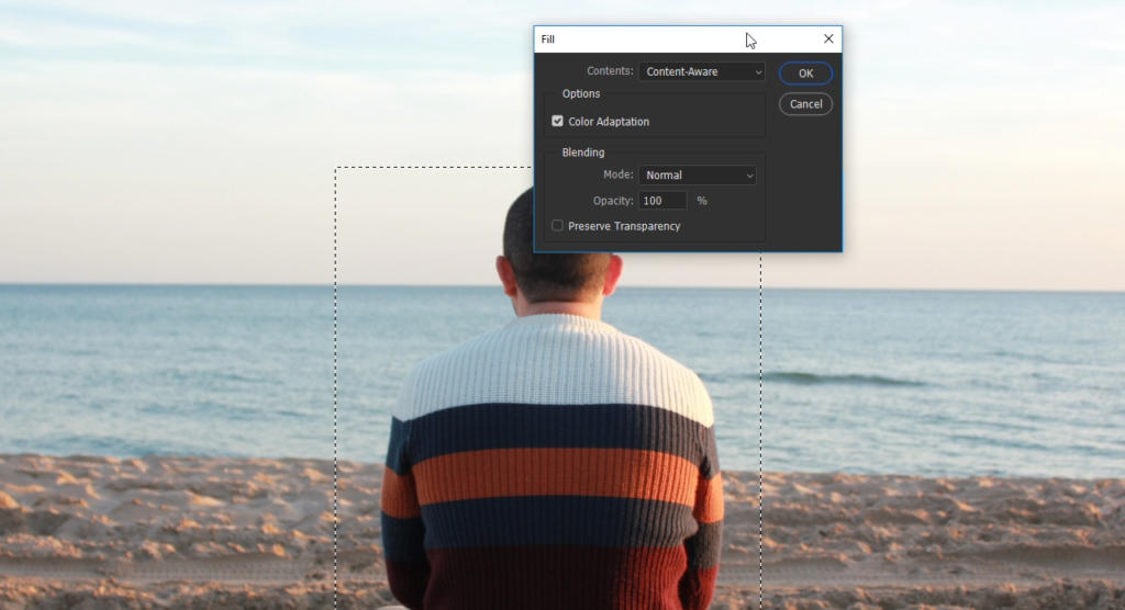Una persona con un maglione a righe è seduta su una spiaggia sabbiosa, di fronte all'oceano. È aperta una finestra di dialogo di Photoshop, che mostra opzioni come sfocatura e riempimento in base al contenuto, che si fondono perfettamente con i colori tenui del cielo limpido.
