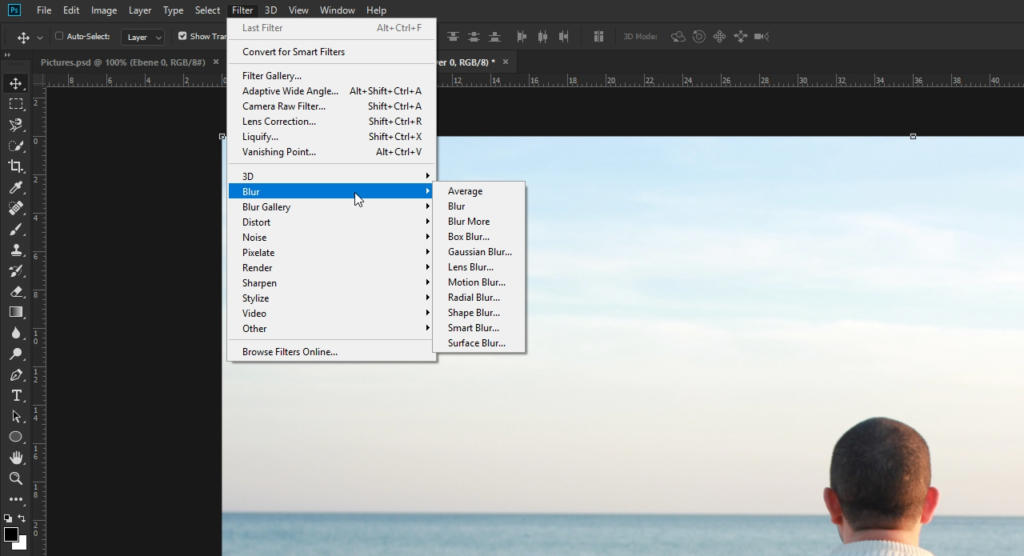 Una persona con i capelli corti sta usando Photoshop per modificare un'immagine sul suo computer. Il menu dei filtri del software, che include opzioni come Sfocatura e Blur More, è aperto. Un mare calmo e un cielo limpido sono visibili nella foto in fase di modifica.