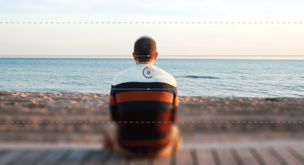 Una persona con un maglione a righe è seduta su una panchina di legno, a guardare la sfocatura dell'oceano al tramonto. L'orizzonte è limpido, con il cielo che sfuma dal blu ai toni caldi vicino all'acqua, creando una scena che sembra essere stata delicatamente toccata da Photoshop.