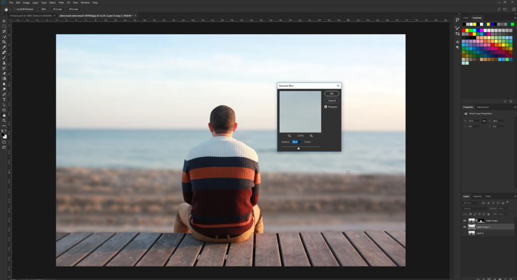 Una persona con i capelli corti, che indossa un maglione a righe, è seduta su un ponte di legno di fronte all'oceano. La scena è sotto il tocco artistico della sfocatura in Photoshop, con una finestra di regolazione Gaussian Blur aperta, che segnala un editing creativo in corso.