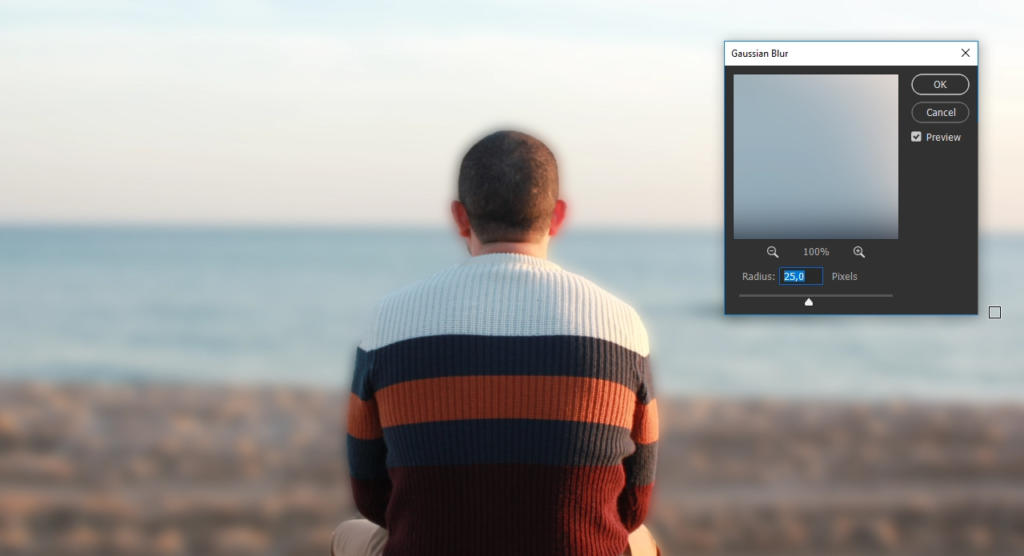 Una persona con un maglione a righe è seduta di fronte all'oceano, con le spalle alla telecamera. In primo piano, è aperta una finestra di modifica della sfocatura digitale in Photoshop, che mostra un cursore di regolazione dei pixel.