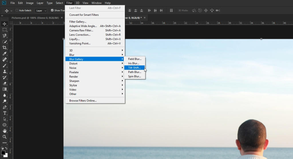 Una persona che modifica meticolosamente una foto di un paesaggio marino in Photoshop, concentrandosi sull'effetto sfocatura. Il menu mostra la scheda Filtro con Blur selezionato e Tilt-Shift evidenziato. Lo sfondo sereno mostra un mare calmo sotto un cielo leggermente nuvoloso.
