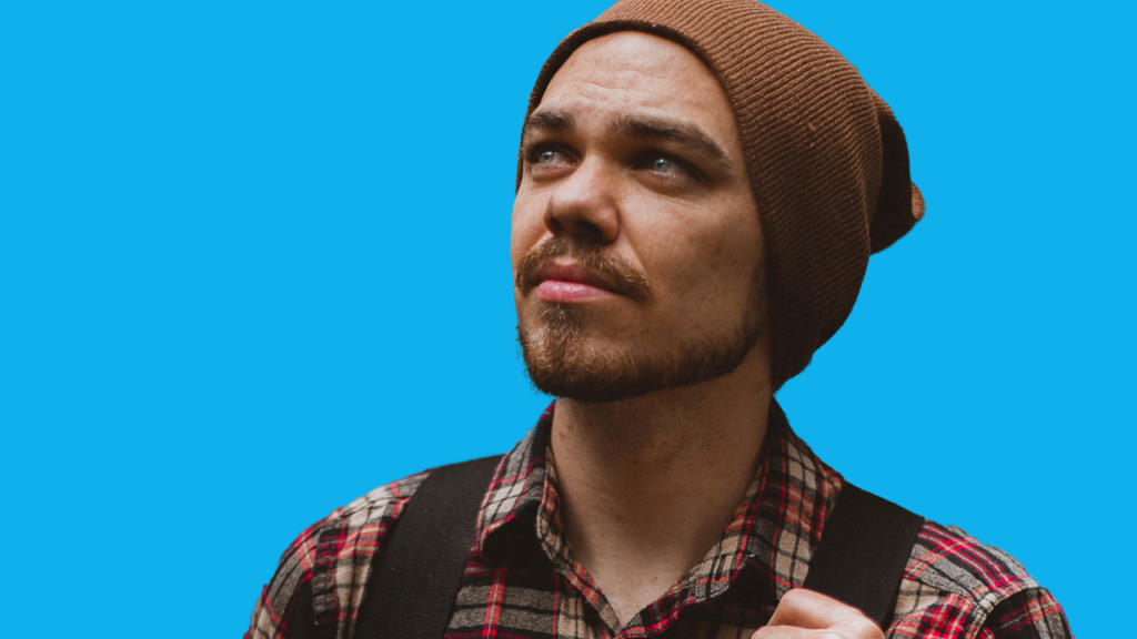 Una persona con la barba e un berretto marrone guarda verso l'alto, indossando una camicia a quadri rossa e grigia e con una tracolla da zaino sulla spalla. Sullo sfondo blu brillante e uniforme, è come se fosse stata ritagliata in Photoshop per dare un'enfasi sorprendente.