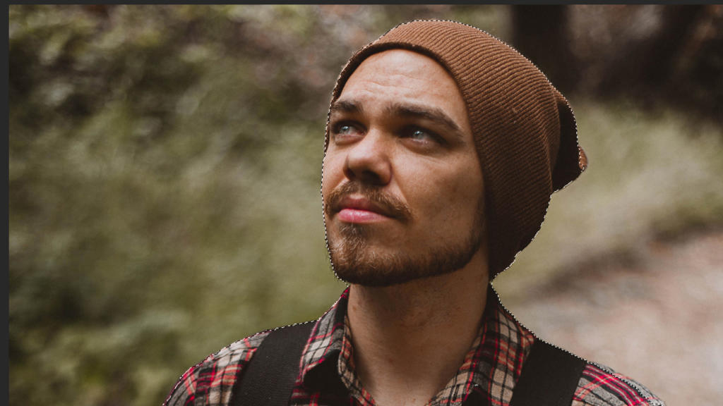 Una persona con la barba, che indossa un berretto marrone e una camicia a quadri, guarda verso l'alto in uno scenario naturale sfocato, come se fosse stato ritagliato alla perfezione con Photoshop.