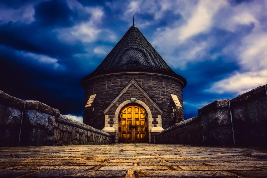Un edificio in pietra con un tetto conico e una porta di legno ad arco è illuminato dall'interno, sullo sfondo di un drammatico cielo serale nuvoloso. Perfetto per essere immortalato con un'app per fotografi, il sentiero di ciottoli che conduce all'ingresso è fiancheggiato da bassi muri in pietra.