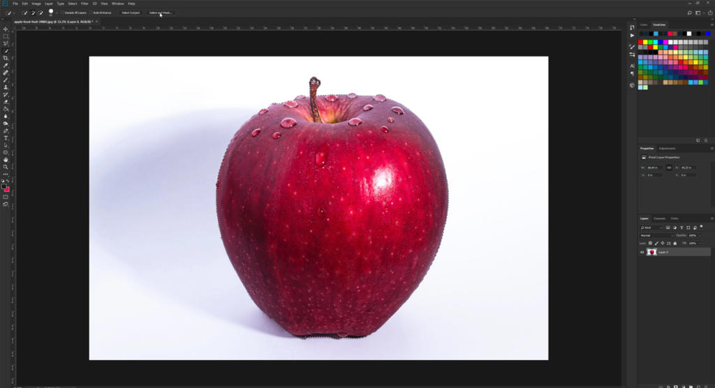 Una mela rossa vibrante, ornata di gocce d'acqua, è appoggiata su uno sfondo bianco. L'immagine è aperta in un software di fotoritocco, dove le barre degli strumenti e le tavolozze dei colori invitano ad attenuare i bordi con Photoshop per una finitura lucida.