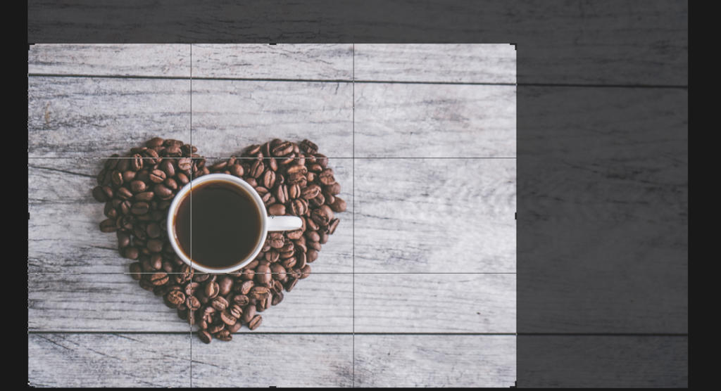 Una tazza di caffè è appoggiata su una superficie di legno, circondata da una disposizione a forma di cuore di chicchi di caffè. La tazza è piena di caffè scuro e la texture rustica in legno grigio sullo sfondo potrebbe ispirare chiunque a ritagliare immagine in Photoshop per il suo prossimo progetto creativo.