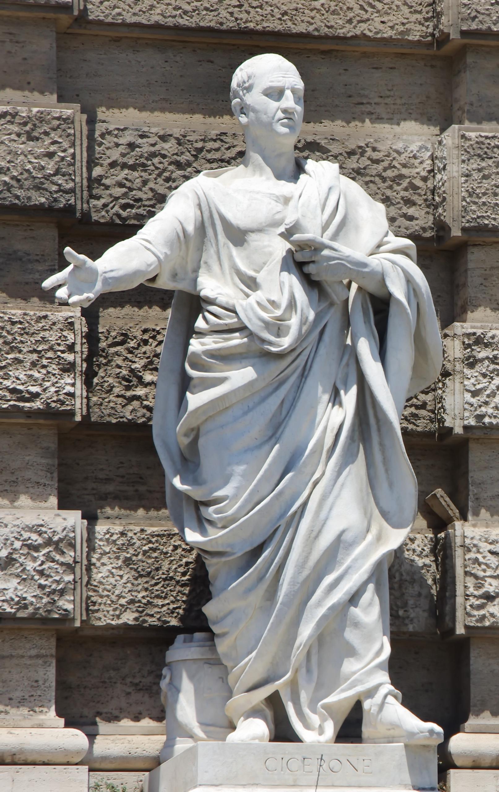 Una statua in marmo bianco di una figura storica in tonaca si erge su un piedistallo con inciso CICERONE. La figura è in posa con un braccio esteso verso l'esterno, su uno sfondo di muro di pietra strutturata.