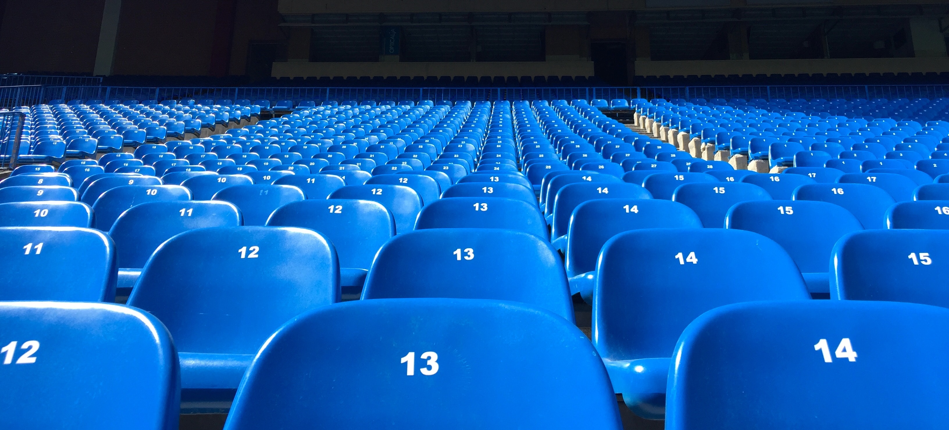 File di posti vuoti blu dello stadio, ognuno numerato in sequenza con numeri bianchi, attendono l'emozione dello Spielplan EM 2021. I posti sono disposti in un lungo schema di fila, creando una vista simmetrica che si estende in lontananza.