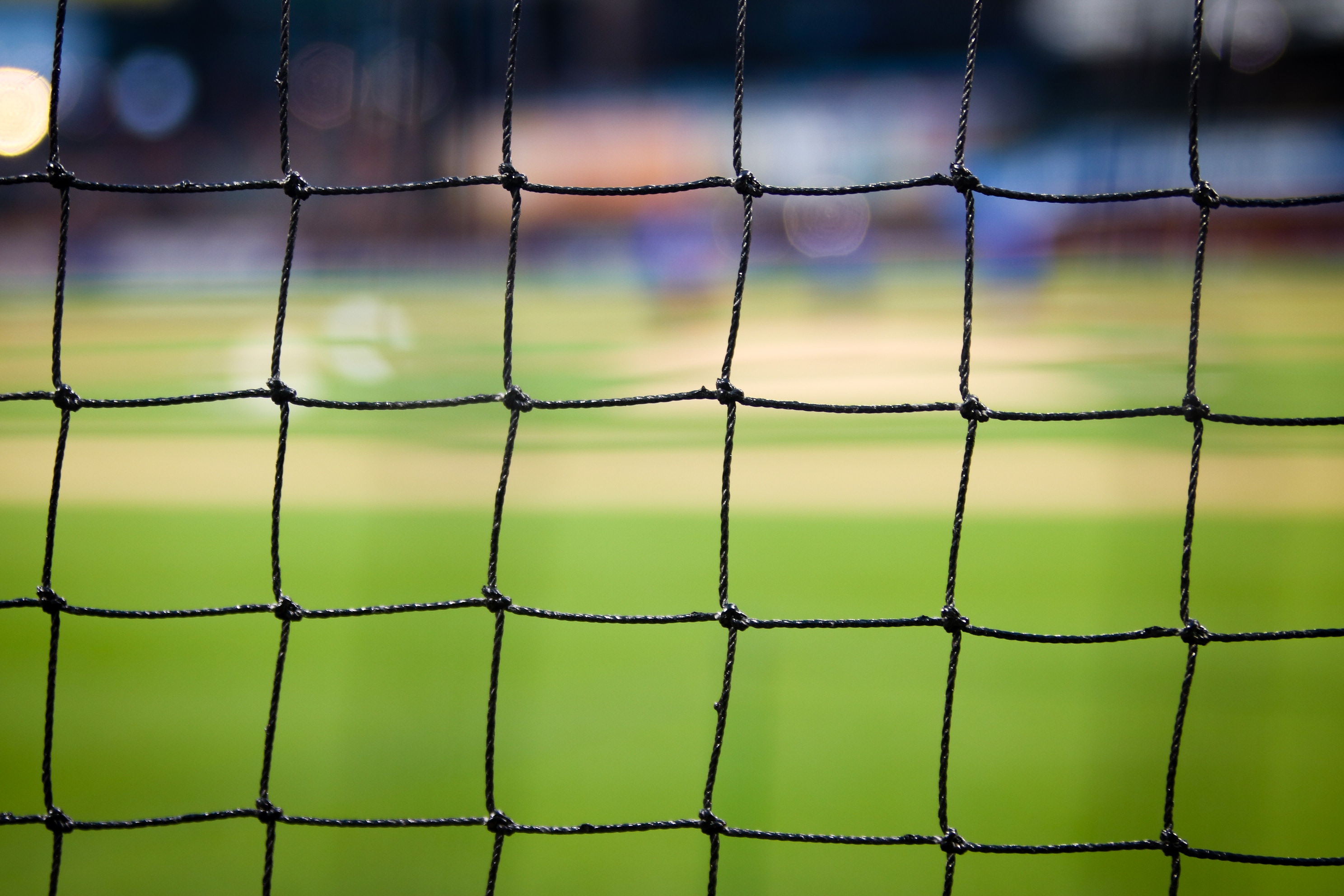 Primo piano di una rete nera con uno sfondo sfocato che mostra un campo sportivo erboso, che ricorda l'eccitazione durante l'EM 2021 Spielplan. Il motivo a griglia della rete è a fuoco, offrendo un sorprendente contrasto con il verde sfocato e gli elementi del campo oltre.