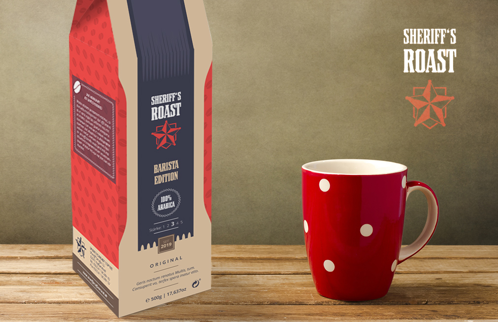 Una confezione di caffè Sheriffs Roast, che mostra il suo Verpackungsdesign, è in piedi su un tavolo di legno accanto a una tazza rossa con pois bianchi. La confezione presenta una sorprendente combinazione di colori rosso e beige con un logo a forma di stella e un testo che la evidenzia come Barista Edition.