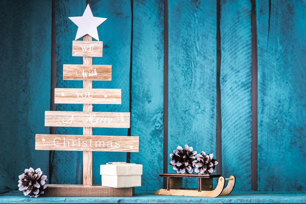 Un albero di Natale in legno con una stella e il messaggio "Ti auguriamo un felice Natale" è affiancato da una piccola scatola regalo e da una slitta in miniatura decorata con pigne, il tutto su uno sfondo rustico in legno blu.