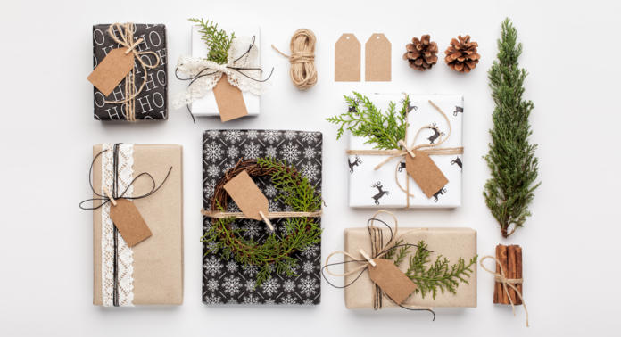 Vari regali incartati con decorazioni naturali come pigne, spago e rametti di verde sono disposti su una superficie bianca. Alcuni regali hanno carta da regalo nera con motivi bianchi, mentre altri usano carta marrone con pizzi e accenti di spago.