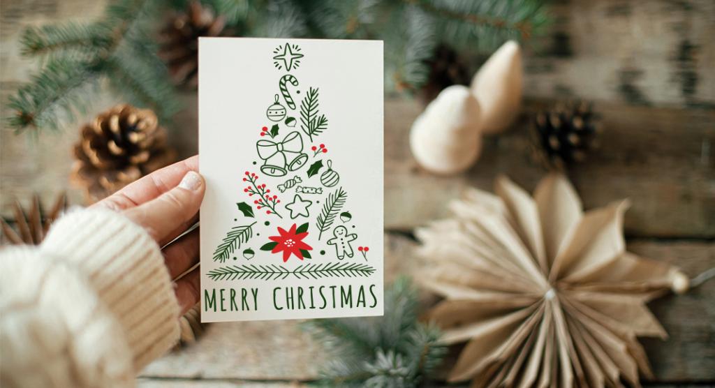Una mano regge un biglietto di auguri natalizio raffigurante un albero fatto di icone festive come un bastoncino di zucchero, un ornamento e una figura di pan di zenzero. Le parole Merry Christmas sono in basso. Lo sfondo include rami di pino, pigne e decorazioni di carta piegate.