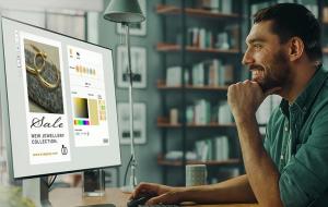 Un uomo sorride allo schermo di un computer che mostra un design per un annuncio pubblicitario di vendita di gioielli. Lo schermo mostra un'immagine di due anelli d'oro e opzioni per colori e caratteri. Sullo sfondo ci sono scaffali con libri e una finestra.