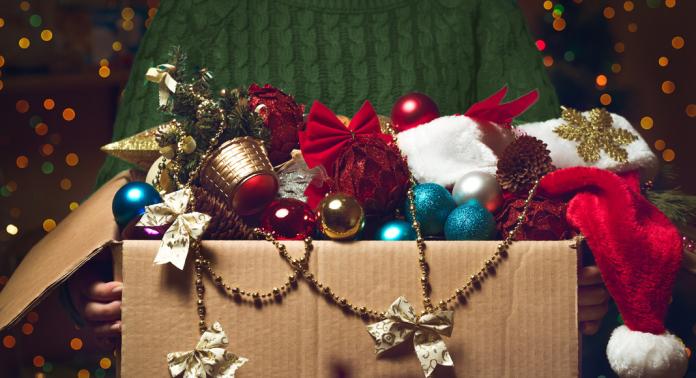 Una persona con un maglione verde tiene in mano una scatola di cartone aperta piena di colorati addobbi natalizi, tra cui palline, pigne, un cappello da Babbo Natale e fiocchi decorativi, con luci festive e sfocate sullo sfondo.