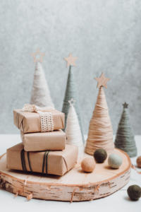 Una composizione festiva presenta piccoli alberi fatti a mano con filato avvolto e stelle in cima. Tre regali ordinatamente incartati sono impilati su una lastra di legno, accompagnati da palline di lana, creando una scena invernale accogliente.