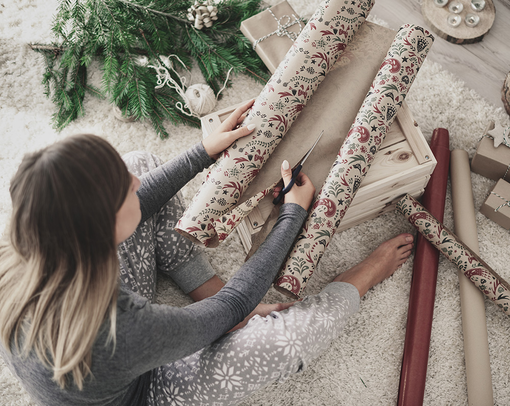 Una persona in un comodo pigiama taglia carta da regalo a tema natalizio su una cassa di legno, circondata da rotoli di carta da regalo e varie piccole scatole regalo. Rami di pino con decorazioni sono sullo sfondo su un tappeto morbido e leggero.