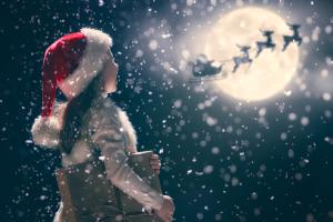 Un bambino che indossa un cappello da Babbo Natale è in piedi nella neve, guardando il cielo mentre tiene in mano un regalo. È visibile la luna piena e le sagome della slitta e delle renne di Babbo Natale volano attraverso di essa.