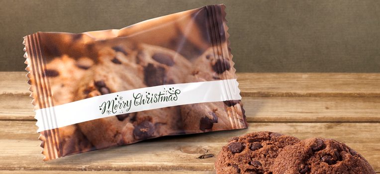 Un pacchetto di biscotti con un'etichetta Merry Christmas è su una superficie di legno. Due biscotti con gocce di cioccolato sono posizionati accanto al pacchetto. Lo sfondo è una parete verde testurizzata.