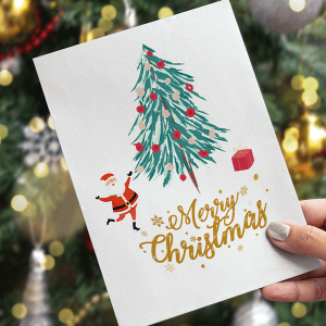 Una mano tiene un biglietto di Natale. Il biglietto raffigura un albero decorato, Babbo Natale e un regalo rosso. Le parole Merry Christmas sono scritte in caratteri dorati. Sullo sfondo c'è un albero di Natale sfocato con ornamenti.