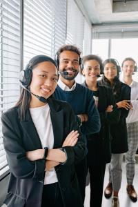 Un gruppo di cinque persone che indossano delle cuffie sono in fila in un ufficio. Sorridono e sembrano rappresentanti del servizio clienti. La stanza luminosa ha grandi finestre con persiane sullo sfondo.